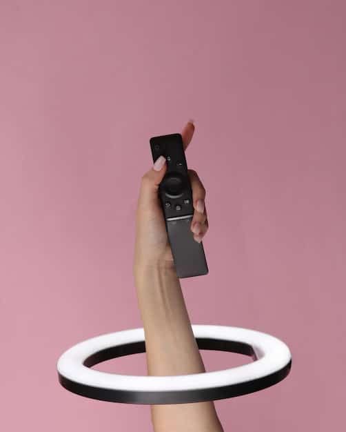 A close-up shot of a hand holding a sleek, modern streaming device remote control, with a blurred television screen displaying a colorful streaming service interface in the background. The focus is sharp on the remote, highlighting its buttons and design.