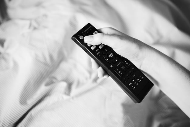 A person's hand holding a remote control, silhouetted against a softly blurred living room with a large screen displaying abstract light patterns, conveying a relaxed and engaging viewing experience at home.