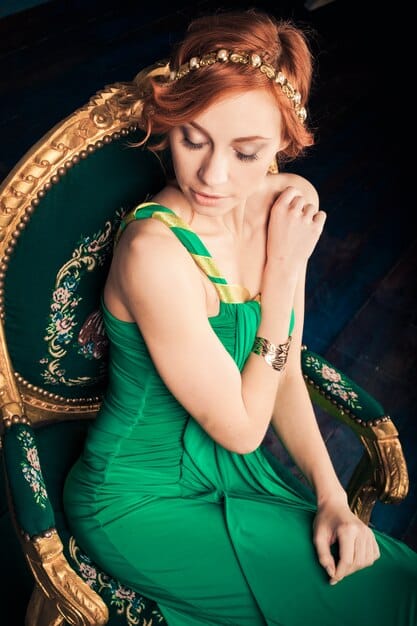 Queen Elizabeth II wearing a dark green dress and a pearl necklace, standing in a grand room with ornate decorations. She has a serious and contemplative expression on her face.